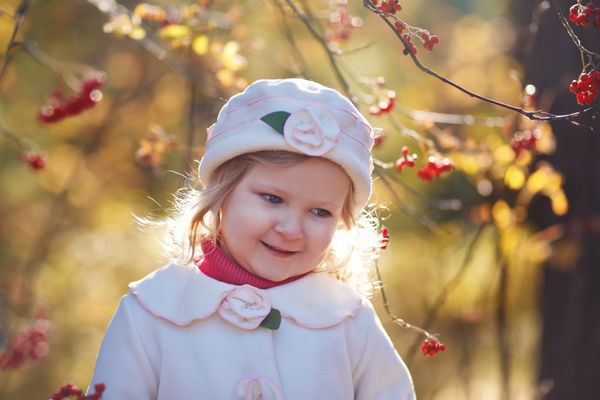 پرتره ای نزدیک از یک دختر کوچک بامزه با کت سفید و کلاهی زیبا که در یک روز آفتابی پاییزی در پارک قدم می زند کودکی که در میان آشبری قرمز ایستاده است