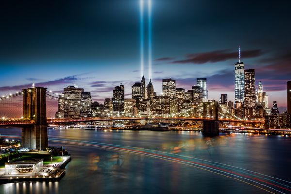 نیویورک - 11 سپتامبر 2014 ادای احترام در یادبود نور پل بروکلین و خط افق پایین منهتن در غروب یک قایق مسیرهای نورانی رنگارنگ را در رودخانه هادسون به جا می گذارد