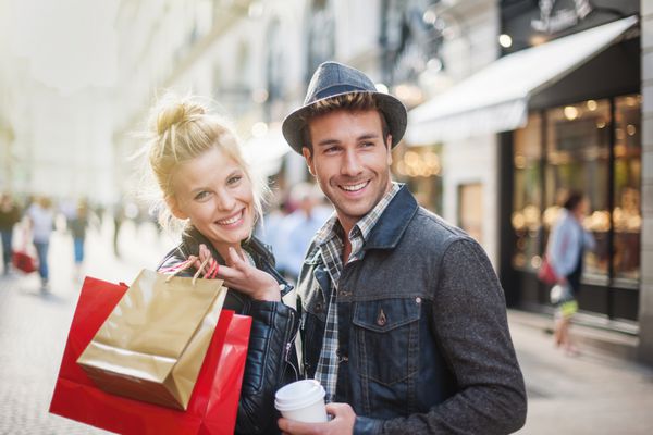 یک زوج جوان شیک پوش در کریسمس در شهر قدم می زنند زن جوان یک ژاکت چرمی می پوشد کیسه های خرید در بازویش است و مرد یک فنجان قهوه در دست دارد