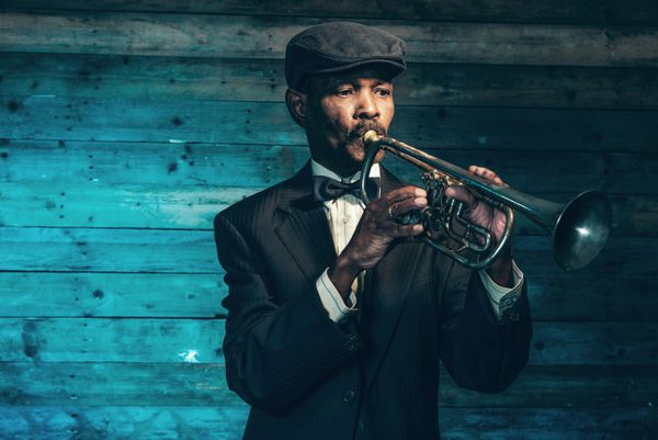 نوازنده قدیمی جاز آفریقایی آمریکایی وینتیج با tpet در مقابل دیوار چوبی قدیمی پوشیدن کت و شلوار مشکی و کلاه