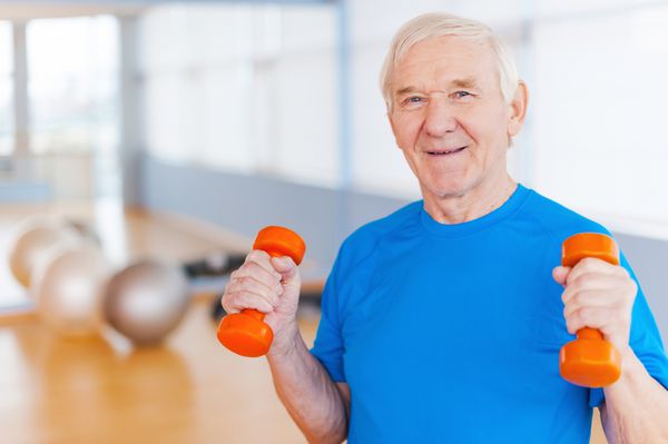 در مسیر بهبودی مرد مسن شادی که با دمبل ورزش می کند و در حالی که داخل خانه ایستاده لبخند می زند