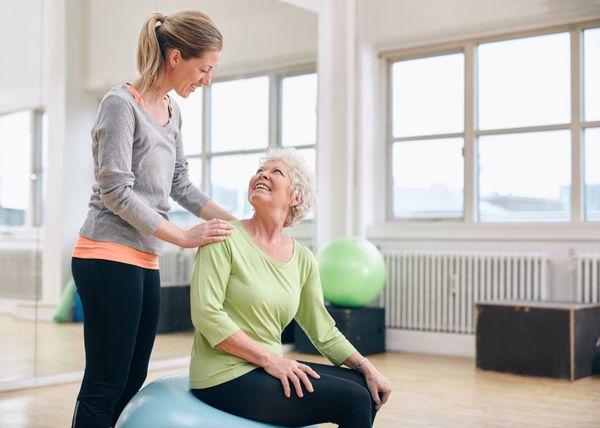 مربی زن به زن سالمند در حال ورزش در باشگاه سلامت کمک می کند زن مسن تر با کمک مربی شخصی در باشگاه