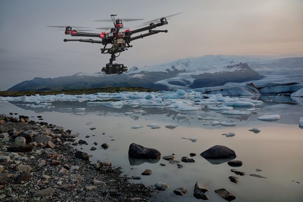 یک پهپاد با ارابه های فرود برآمده و دوربینی که بر فراز کوه های یخ با یک یخچال طبیعی در پس زمینه پرواز می کند