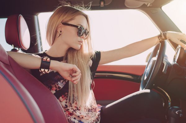 دختر جوانی روی فرمان ماشین اسپرت با داخل قرمز با عینک آفتابی مشکی و بازوهای چرمی مشکی با درج‌های فلزی که به پهلو می‌نشیند و به جاده نگاه می‌کند