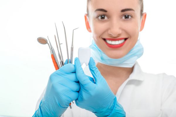 پزشک زن جوانی که تجهیزات دندانپزشکی را در دست دارد و به دوربین در پس زمینه سفید نگاه می کند