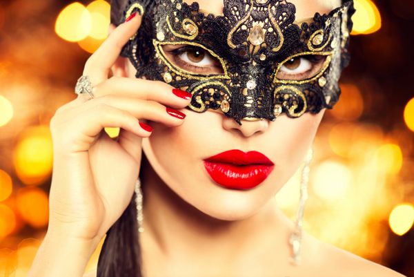 زن مدل زیبایی با ماسک کارناوال بالماسکه ونیزی در مهمانی روی پس زمینه تیره تعطیلات جشن کریسمس و سال نو دختری با آرایش و مانیکور تعطیلات لب و ناخن قرمز