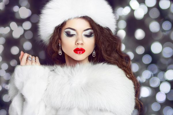 مدل دختر مد با کت خز و کلاه خزدار سفید زن زیبای زمستانی با لباس های لوکس بر روی پس زمینه چراغ های کریسمس بوکه