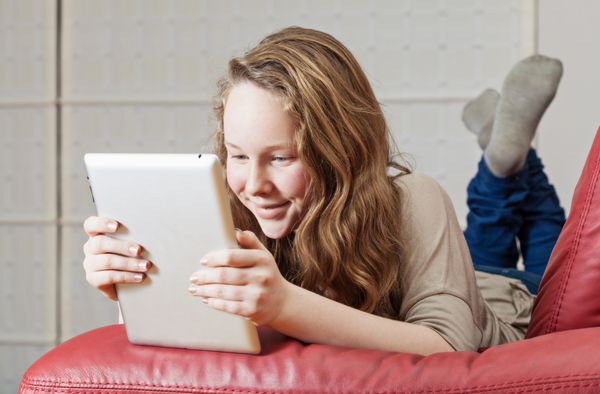 دختر نوجوان خندان که روی مبل با رایانه لوحی دراز کشیده است