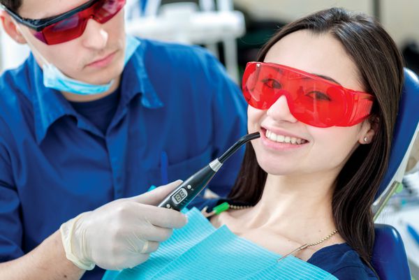 دندانپزشک بیمار را درمان می کند بیمار زن خندان که روی صندلی دندانپزشکی با عینک UV نشسته و مستقیماً به دوربین لبخند می‌زند در حالی که یک لامپ UV دندانپزشکی را نزدیک دهان خود نگه داشته است