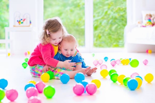 دو کودک کوچک شاد دختر نوپای فرفری بامزه و یک نوزاد پسر بامزه همراه با توپ های رنگارنگ در یک اتاق آفتابی سفید با پنجره بزرگ