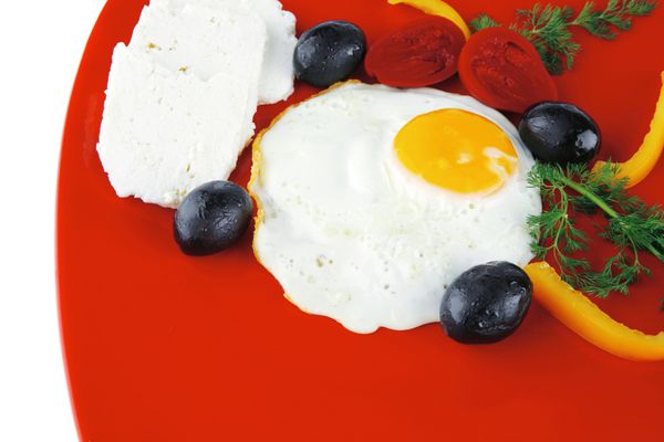 چشم تخم مرغ سوخاری سرخ شده با پنیر فتا سفید بز روی بشقاب قرمز جدا شده روی پس زمینه سفید با زیتون سیاه و سبزیجات