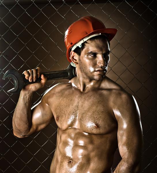 مرد کارگر عضلانی زیبایی با کلاه ایمنی با آچار بزرگ در دست روی زمینه حصار توری