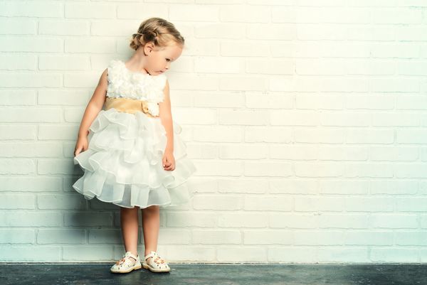 دختر بچه فرشته ای با لباس سفید زیبا کنار دیوار آجری سفید ایستاده است دوران کودکی