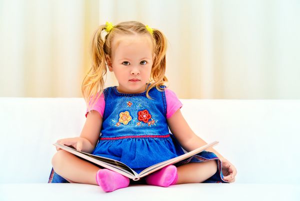 دختر کوچک زیبایی که روی مبل نشسته و به کتاب مصور کودکان نگاه می کند کودکی شاد