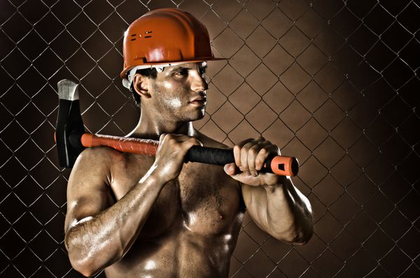 مرد هلی کوپتر کارگر خسته عضلانی با کلاه ایمنی با تبر بزرگ سنگین در دست روی پس زمینه حصار توری