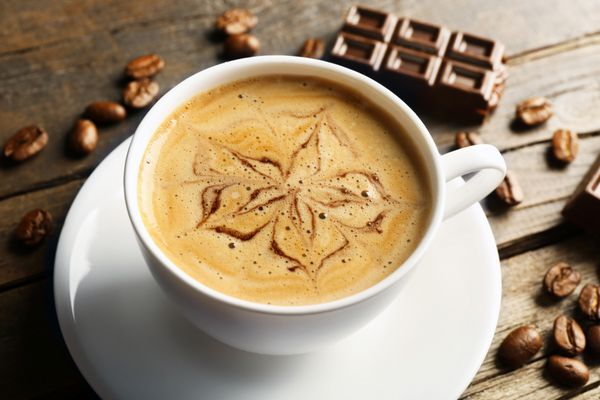 فنجان قهوه لاته آرت با دانه ها و شکلات روی میز چوبی نمای نزدیک