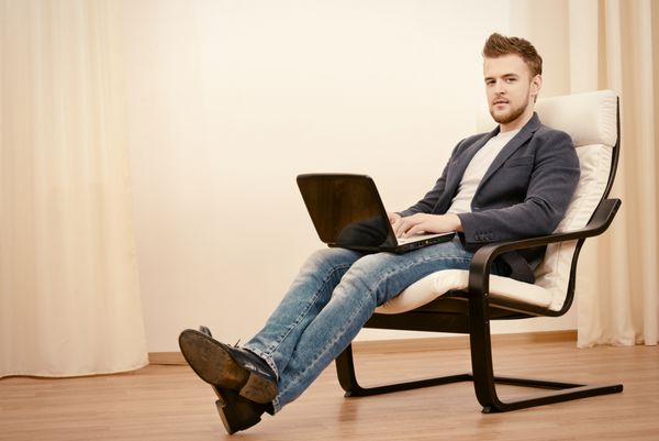مرد جوان خوشحال با استفاده از لپ تاپ که روی صندلی راحتی در خانه نشسته است