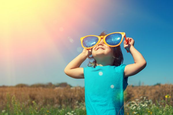 دختر کوچولوی شاد با عینک آفتابی بزرگ که در چمنزار قدم می زند و به خورشید نگاه می کند
