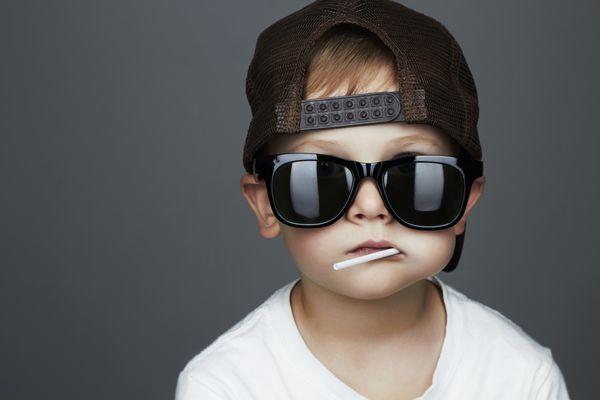 پسر جوان خنده دار در حال خوردن آبنبات چوبی کودک شیک در عینک آفتابی