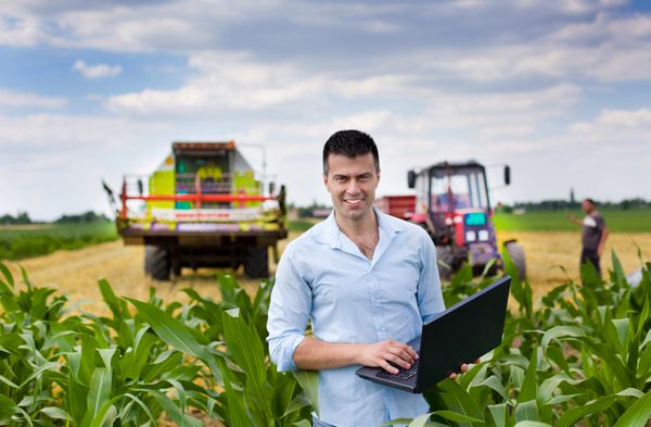 کشاورز جذاب جوان با لپ تاپ ایستاده در مزرعه ذرت تراکتور و کمباین کار در زمینه گندم در پس زمینه