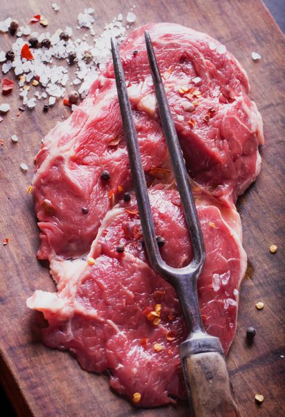 گوشت تازه خام ریبای استیک با چاشنی و چنگال گوشت روی تخته چوبی قدیمی