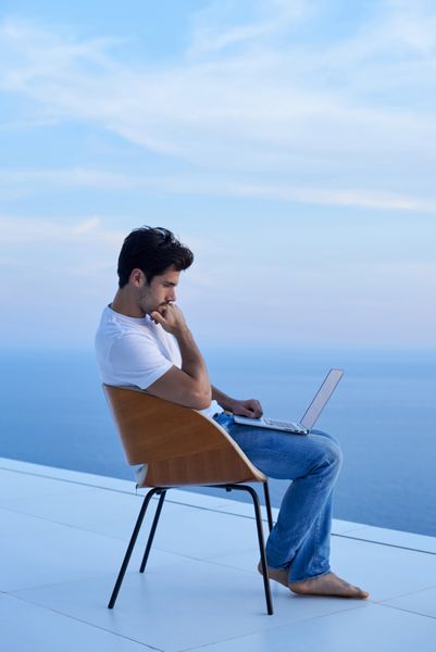 جوان زیبا در حال استراحت و کار با رایانه لپ تاپ در بالکن خانه در حالی که به غروب آفتاب نگاه می کند