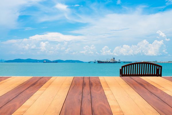 میز خالی تخته چوبی قدیمی در مقابل چشم انداز تابستانی کف چوبی پرسپکتیو روی دریا با نیمکت چوبی - می تواند برای نمایش یا مونتاژ محصولات شما استفاده شود نمای رو به پس زمینه دریا