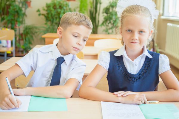 دانش‌آموزان یا همکلاسی‌های کلاس درس با هم پشت میز نشسته‌اند پسر امتحان دختر را می نویسد او به دفترچه او نگاه می کند آنها لباس مدرسه پوشیده اند روی میز دفتر و خودکار وجود دارد