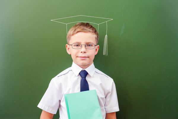 پسر مدرسه ای موفقی که در نزدیکی تخته سیاه در کلاس درس ایستاده است با لباس مدرسه کلاه با گچ و عینک آفتابی تصویر شده