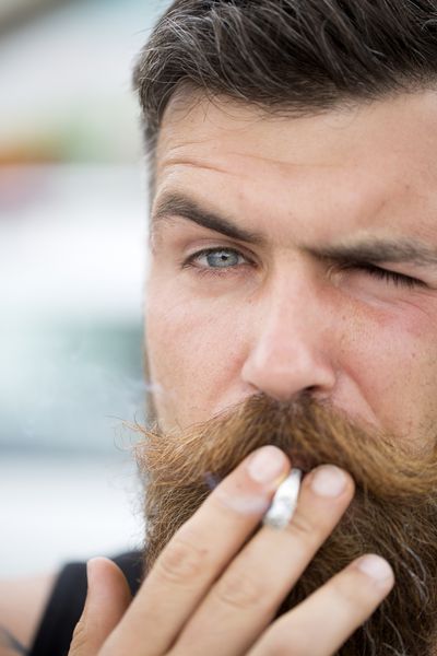 پرتره مرد جذاب نتراشیده با ریش و دسته سبیل در حال سیگار کشیدن به جلو با نمای نزدیک در فضای باز تصویر عمودی