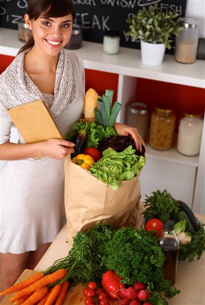 زن جوانی که کیسه خرید مواد غذایی با سبزیجات در آشپزخانه ایستاده است