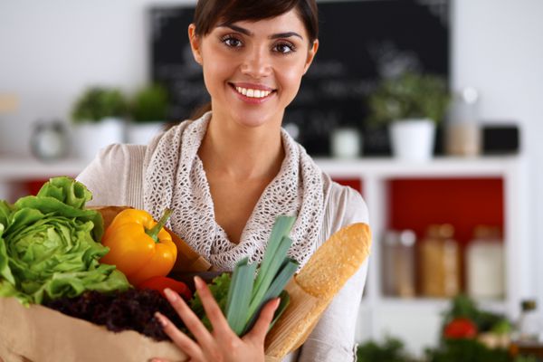زن جوانی که کیسه خرید مواد غذایی با سبزیجات در آشپزخانه ایستاده است