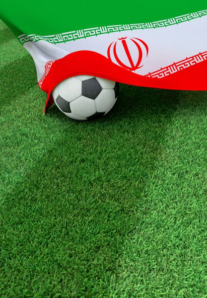 توپ فوتبال و پرچم ملی ایران بر روی چمن سبز قرار دارد
