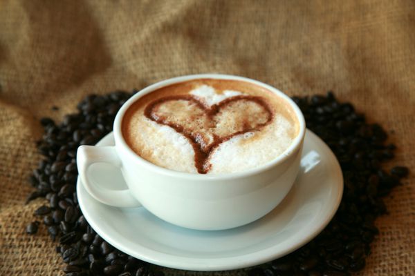 قهوه برای دوستداران قهوه یک فنجان قهوه یا اسپرسو با قلب در فوم که در بستری از دانه‌های قهوه آسیاب نشده روی پس‌زمینه کرباسی قرار گرفته است
