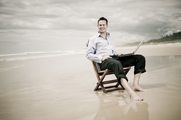 مرد تجاری با لپ تاپ روی یک صندلی در ساحل نشسته است تصویر رنگی گرم با قهوه ای