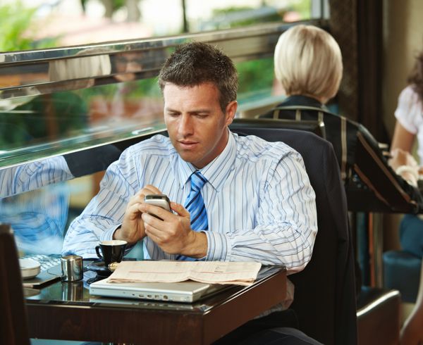 تاجری که با تلفن همراه پشت میز کافه نشسته است زنان جوانی که در پس زمینه شیرینی دارند