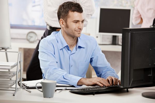 تاجری که در دفتر کار می کند پشت میز می نشیند روی صفحه کلید تایپ می کند به صفحه کامپیوتر نگاه می کند