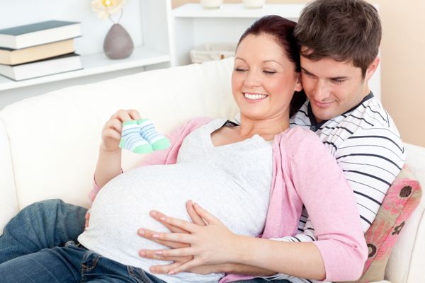 پرتره یک زن باردار شاد که کفش های بچه را در دست دارد و همسرش روی مبل خانه