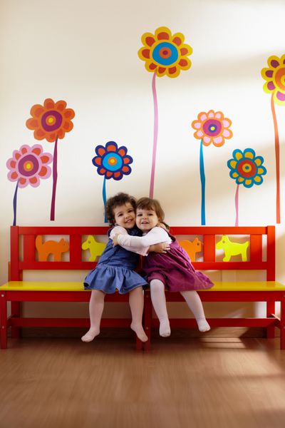 دختران پیش دبستانی قفقازی و اسپانیایی تبار در مهدکودک روی نیمکت نشسته و لبخند می زنند شکل عمودی تمام طول نمای جلو