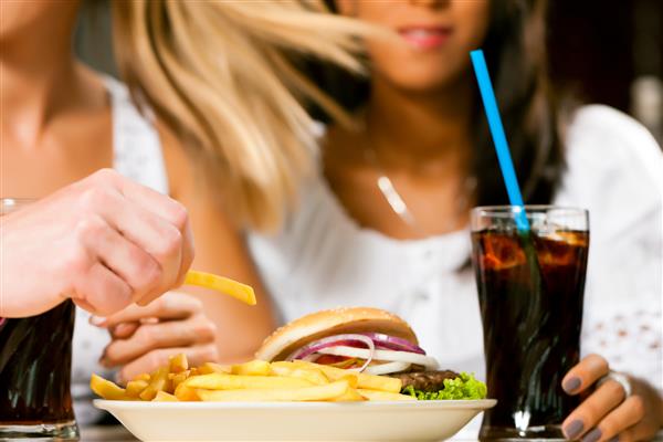 دو زن - یکی آفریقایی آمریکایی - در حال خوردن همبرگر و نوشیدن نوشابه در یک غذاخوری فست فود روی غذا تمرکز کنید