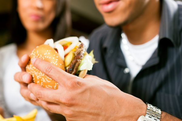 مرد خوشحال آمریکایی آفریقایی تبار در یک رستوران فست فود در حال خوردن همبرگر با دوست دخترش