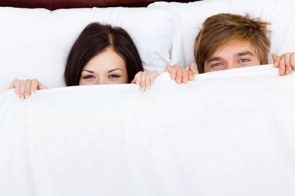 زوج جوان دوست‌داشتنی که روی تخت دراز کشیده‌اند لبخند شادی که به دوربین نگاه می‌کنند پوشش زیر پتو زاویه بالا نمای بالا