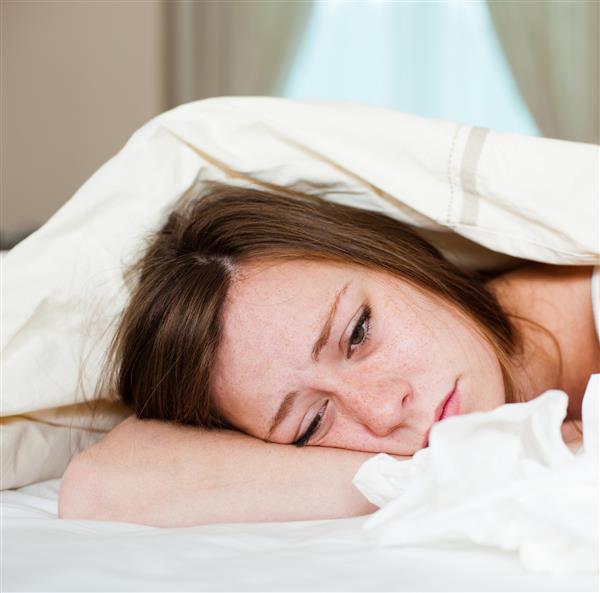 یک خانم جوان بیمار در رختخواب با سرش زیر پتو و انبوهی از دستمال کاغذی در مقابلش