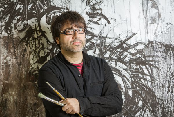 هنرمند نقاش با عینک سیاه که در مقابل نقاشی دیواری خود ژست گرفته است