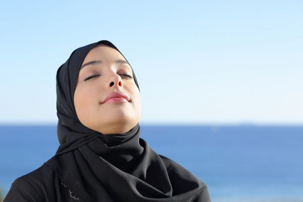 زن عرب سعودی در حال تنفس عمیق هوای تازه در ساحل با اقیانوس و افق در پس زمینه