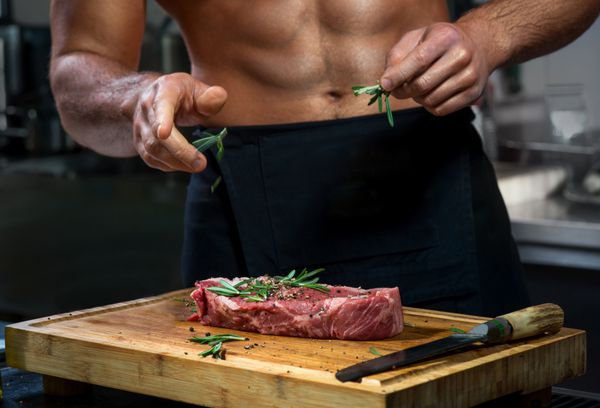 آشپز با بدن در حال پختن یا تزئین گوشت خام با رزماری در آشپزخانه رستوران