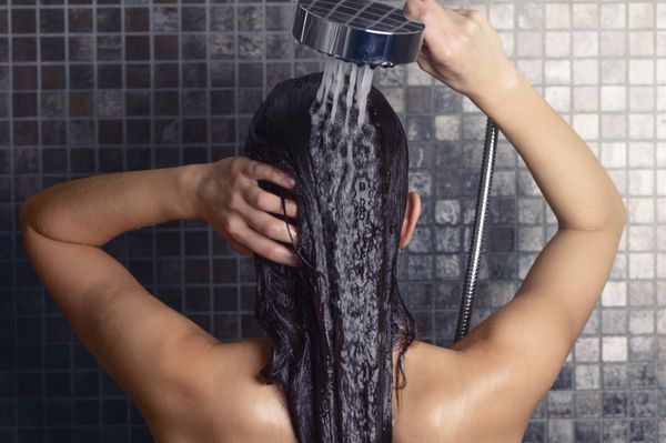زن جوان در حال شستن موهای بلندش زیر دوش ایستاده و پشتش به دوربین و در حالی که سرش تا حدی به پهلو چرخیده روی کاشی‌های موزاییک خاکستری آن‌ها را زیر فواره آب می‌شوید