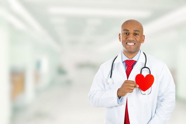 دکتر خندان خوشحال که قلب قرمز را در دست دارد و در راهروی بیمارستان در پس زمینه مطب کلینیک ایستاده است قرار ملاقات مراقبت های قلب و عروق و مفهوم بررسی