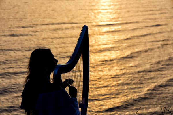 زن سیلوئت در هنگام غروب آفتاب در سانتورینی یونان در کنار دریا چنگ می نوازد