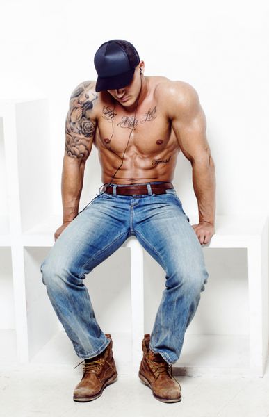 مرد عضلانی بدون پیراهن با خالکوبی با شلوار جین آبی که روی سکوی مربعی روی دیوار سفید نشسته است